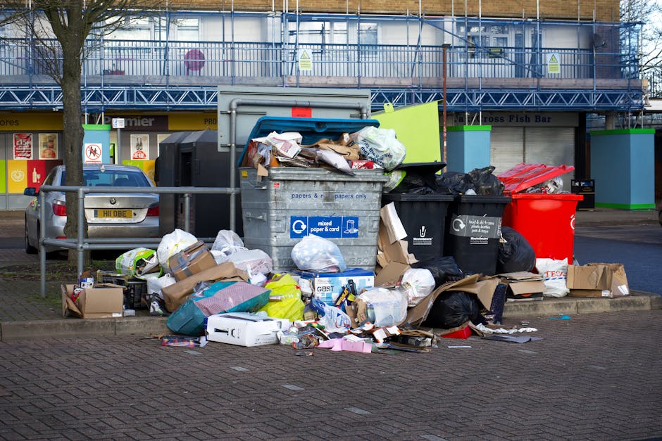 A large pile of assorted waste and rubbish bags on a paved sidewalk area in front of a commercial building with blue scaffolding. The waste includes black plastic rubbish bags, cardboard boxes, flattened cartons, plastic packaging, and loose paper debris. Some items are spilling out of a grey mixed paper and cardboard bin labeled 'papers & cards,' while others are stacked or placed nearby. To the left, a small silver car is parked behind a metal railing, partly obscuring the waste. The background features a storefront with signs and a green and blue colour scheme, including a sign for 'Premier' and an entrance to a fish bar. The scene is set during daylight with natural lighting, showing an environment that appears to be a public space or parking area often associated with private rubbish collection and waste management services, such as those provided by Waste Disposal Ruislip for alternative disposal or on-site clearance.