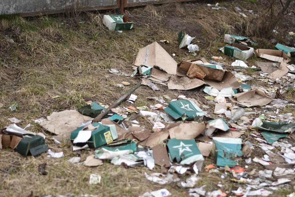 A large pile of assorted waste and rubbish bags on a paved sidewalk area in front of a commercial building with blue scaffolding. The waste includes black plastic rubbish bags, cardboard boxes, flattened cartons, plastic packaging, and loose paper debris. Some items are spilling out of a grey mixed paper and cardboard bin labeled 'papers & cards,' while others are stacked or placed nearby. To the left, a small silver car is parked behind a metal railing, partly obscuring the waste. The background features a storefront with signs and a green and blue colour scheme, including a sign for 'Premier' and an entrance to a fish bar. The scene is set during daylight with natural lighting, showing an environment that appears to be a public space or parking area often associated with private rubbish collection and waste management services, such as those provided by Waste Disposal Ruislip for alternative disposal or on-site clearance.