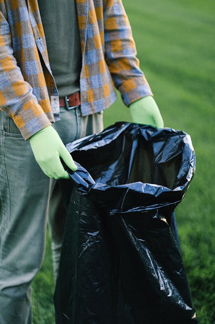Close-up of a person wearing a checkered yellow, grey, and blue shirt over a grey T-shirt, with olive green gloves on their hands, holding open a large black plastic rubbish bag. The background shows a well-maintained grassy area, suggesting an outdoor setting such as a garden or park. The person's lower body, including khaki pants and a brown belt, is partially visible. The black rubbish bag appears partially filled, ready for waste disposal or collection, consistent with private waste removal or on-site clearance activities. This scene reflects a waste management process, where an individual prepares rubbish for transportation, managed by a waste disposal company like Waste Disposal Ruislip, offering alternative waste handling services outside traditional council collections.