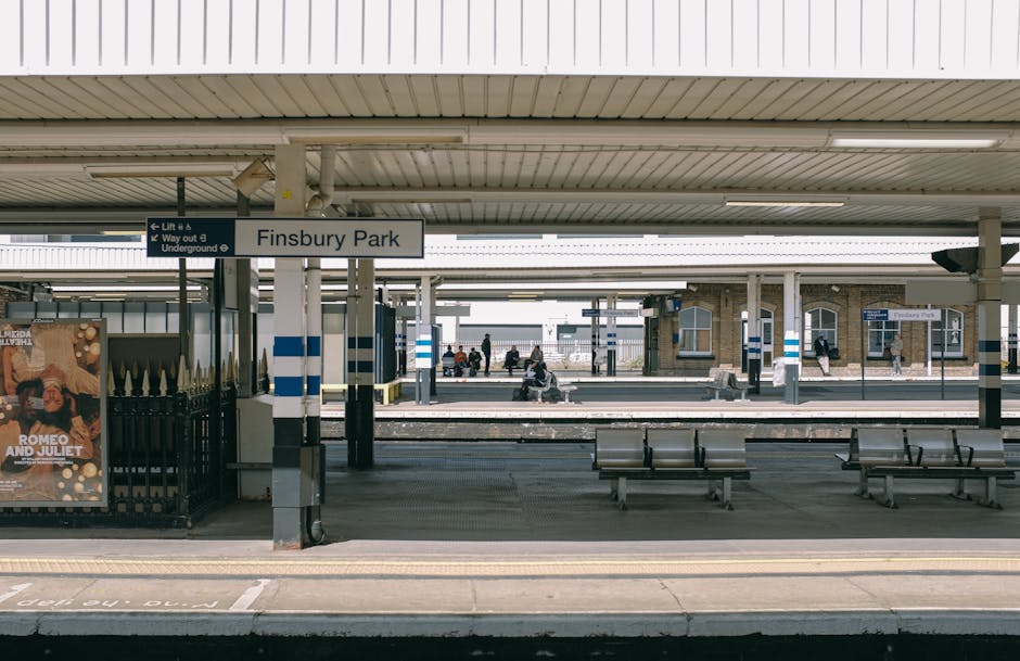 The image depicts Finsbury Park train station platform with a shelter overhead made of metallic panels painted in light grey. In the foreground, a sign displays the station name 'Finsbury Park' in black text on a white background, mounted on a metal pole. The platform surface is concrete with tactile paving strips along the edge for safety. Several metal benches with wooden slats are arranged along the platform, some occupied by waiting passengers. To the left, a black metal gate with an advertising poster for a theatrical production is visible, featuring warm tones and a partially obscured image of a performer. Further back, railway tracks run parallel to the platform, with a few passengers standing or sitting on benches, some waiting for trains. The background reveals additional platforms and a building with classic brick architecture, large windows, and a small shelter. The scene is illuminated by natural daylight, under an overcast sky, capturing a typical busy urban railway station setting associated with public transportation hubs, distinct from private waste or rubbish removal services, yet relevant to transportation infrastructure supporting urban waste management needs.